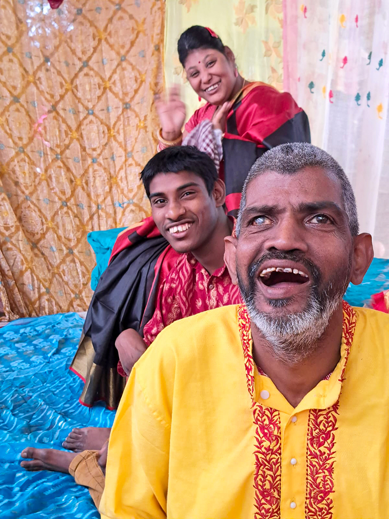 Older man with beard and gray hair laughing wearing a yellow kurta with red trim. Behind him is a younger man smiling in a fancy red shirt and a young smiling woman in a pink and black sari waving.