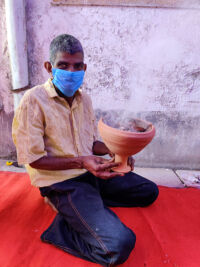 shuktara - man wearing mask holding a medium size unglazed clay pot with smoke rising off the top