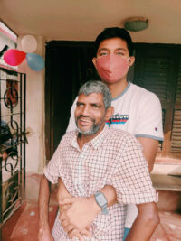 shuktara - man with gray hair sitting, young man in pink mask standing behind him with his arms around the seated man's waist