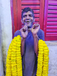 shuktara - man laughing in front of red painted shutters holding bright yellow marigold garlands
